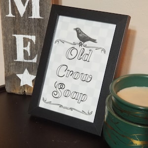 May include: A framed print with the words "Old Crow Soap" and a crow illustration. The print is in a black frame and sits on a dark surface. A wooden sign with white letters and a star is visible.