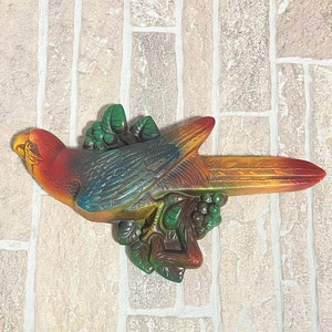May include: A colorful ceramic parrot figurine with a red, orange, yellow, green, and blue paint scheme. The parrot is perched on a branch with green leaves and berries.