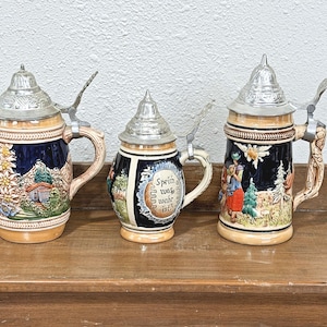 May include: Three ceramic beer steins with metal lids and handles. The left stein features a mountain landscape. The center stein displays the text "Sprich was wahr ist" in a decorative frame. The right stein depicts a figure in a field.
