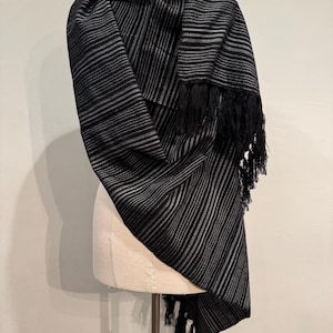 May include: A black and gray striped scarf with fringe detail. The scarf is draped over a mannequin, showcasing the woven pattern and texture. The scarf's design features alternating black and gray stripes.