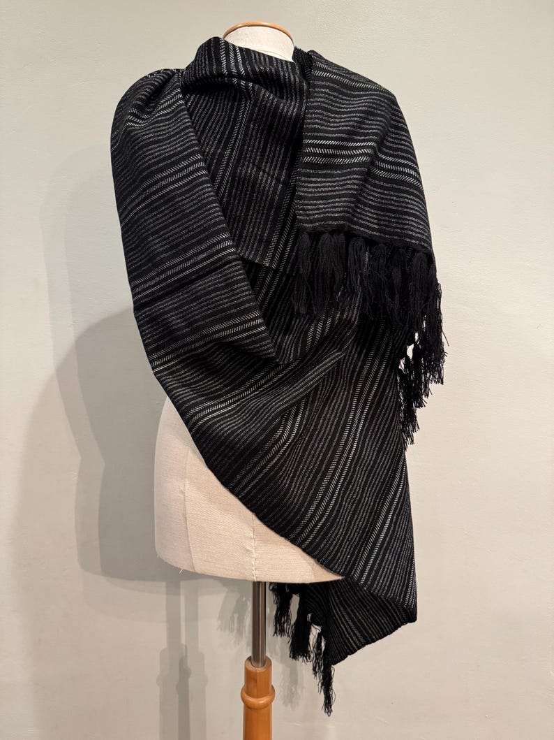 May include: A black and gray striped scarf with fringe detail. The scarf is draped over a mannequin, showcasing the woven pattern and texture. The scarf's design features alternating black and gray stripes.