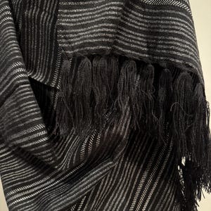 May include: Black and white striped scarf with black fringe. The scarf features a repeating pattern of thin white stripes against a black background. The fringe is thick and hangs from the bottom edge of the scarf.