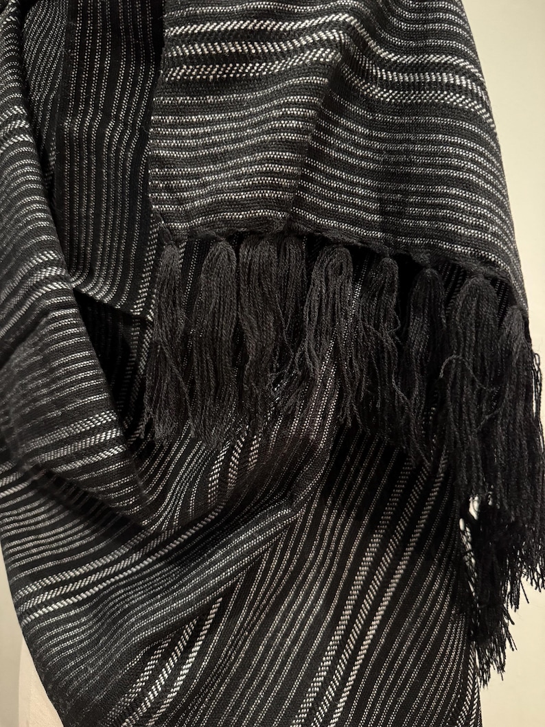 May include: Black and white striped scarf with black fringe. The scarf features a repeating pattern of thin white stripes against a black background. The fringe is thick and hangs from the bottom edge of the scarf.