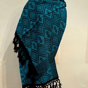 Puede incluir: Un chal tejido en color turquesa y negro con un patrón geométrico de diamantes y detalles florales. El chal tiene flecos negros en el borde inferior. El textil está drapeado sobre un maniquí.