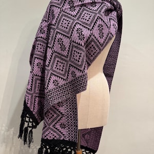 Puede incluir: Un chal tejido en tonos lavanda y negro, drapeado sobre un maniquí. El chal presenta un patrón geométrico de diamantes con detalles florales. Flecos negros adornan los bordes, agregando textura y detalle.