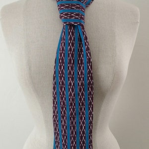 May include: A patterned necktie displayed on a mannequin. The tie features a repeating pattern of teal vertical stripes and a woven design in shades of purple, white, and teal. The tie is knotted at the neck.