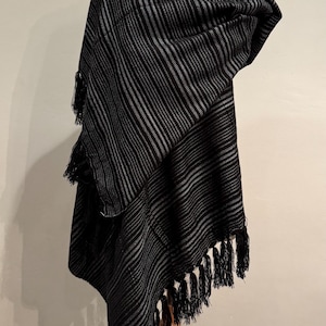 May include: A black and gray striped shawl with fringe. The shawl is draped over a wooden mannequin. The fabric has a textured appearance with vertical stripes. The fringe hangs along the bottom edge.