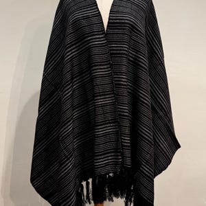 May include: A black and gray striped poncho with a V-neck design. The poncho has a fringe trim along the bottom edge and is displayed on a mannequin. The fabric appears to be a soft, woven material.