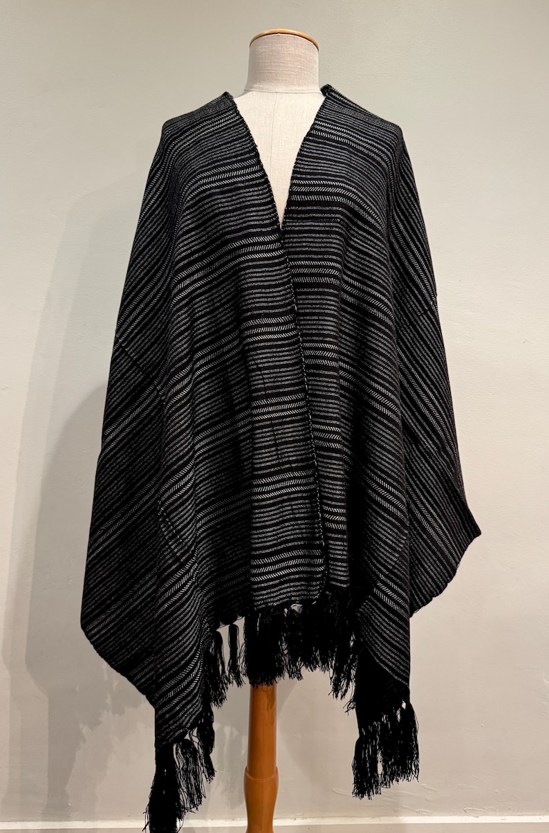 May include: A black and gray striped poncho with a V-neck design. The poncho has a fringe trim along the bottom edge and is displayed on a mannequin. The fabric appears to be a soft, woven material.