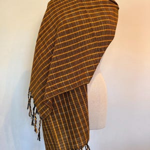 Peut inclure: Écharpe à carreaux marron et or avec franges. L'écharpe est drapée sur un mannequin. Le motif à carreaux présente de fines lignes dorées sur un fond marron foncé. L'écharpe a une frange de fils torsadés aux extrémités.