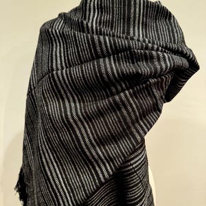May include: A black and silver striped shawl draped over a mannequin. The shawl has vertical stripes of varying widths, creating a textured appearance. The fabric appears soft and drapes elegantly, with fringed edges.