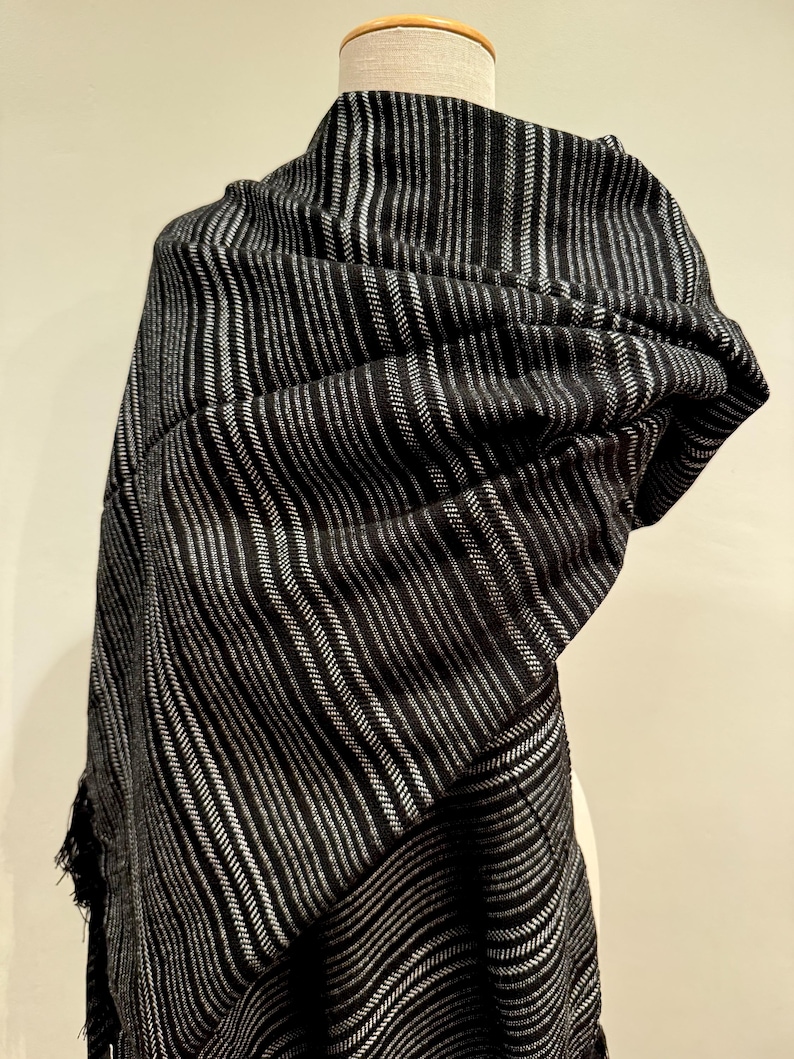 May include: A black and silver striped shawl draped over a mannequin. The shawl has vertical stripes of varying widths, creating a textured appearance. The fabric appears soft and drapes elegantly, with fringed edges.