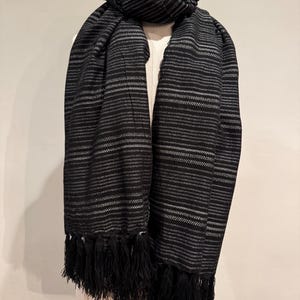 May include: A black and white striped scarf with a fringe. The scarf is draped over a mannequin. The scarf has horizontal stripes with a fringe at the bottom. The scarf is made of a soft material.