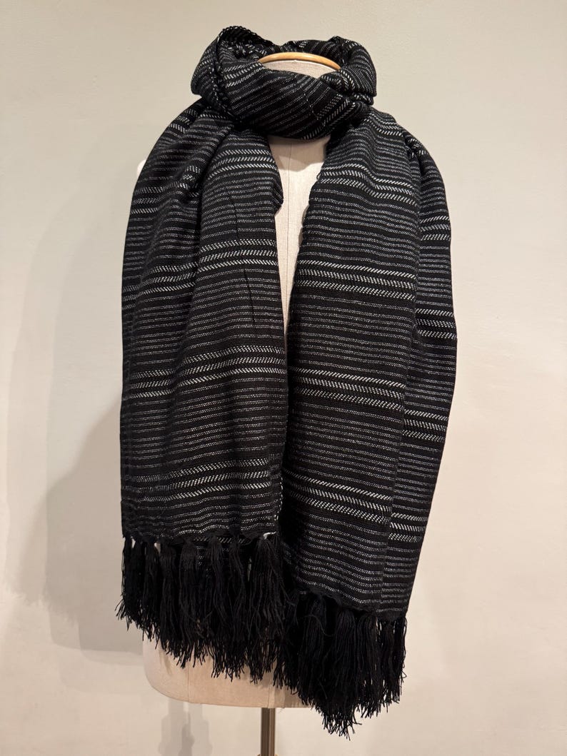 May include: A black and white striped scarf with a fringe. The scarf is draped over a mannequin. The scarf has horizontal stripes with a fringe at the bottom. The scarf is made of a soft material.