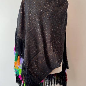 May include: A black shawl with gold flecks and colorful fringe. The shawl is draped over a mannequin, showcasing its texture and design. The fringe features vibrant hues of pink, green, orange, and purple, adding a pop of color.