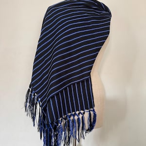 May include: A black shawl featuring thin, vertical blue and white pinstripes throughout the fabric. The garment is draped over a mannequin and finished with long, knotted black and blue fringe tassels along the bottom edge of the textile.