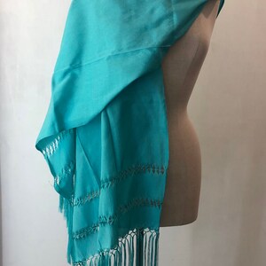 May include: A turquoise shawl with a delicate lace pattern and fringe. The shawl is draped over a dress form.