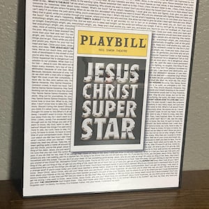 May include: A framed Playbill for the musical "Jesus Christ Superstar" with a silver title. The background is a collage of text, and the Playbill is set against a brown surface.