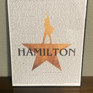 Puede incluir: Impresión enmarcada con la palabra "HAMILTON" en letras naranjas desgastadas y una silueta. El fondo está lleno de texto pequeño, probablemente letras del musical.