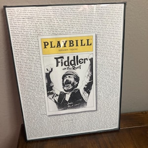 May include: A framed playbill for the musical "Fiddler on the Roof" at the Gershwin Theatre. The playbill features a black and white photo of the actor playing Tevye, with his arms raised in the air. The playbill is surrounded by a black frame.