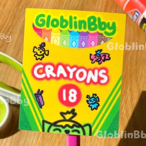 May include: A yellow crayon box with the text "CRAYONS 18" and the brand name "GloblinBby". The box is held by a pink pencil. Other items in the image include scissors, a glue stick, and a ruler.