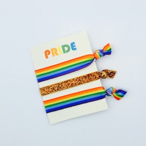 Pode incluir: Três elásticos de cabelo arco-íris com a palavra "PRIDE" impressa em um cartão branco. Os elásticos de cabelo são feitos de elástico e têm um acabamento brilhante.