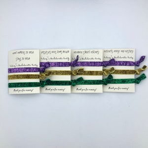 Peut inclure: Quatre cartes avec des dictons diff&eacute;rents li&eacute;s &agrave; la Nouvelle-Orl&eacute;ans, chacune avec trois &eacute;lastiques pour cheveux en violet, or et vert. Les dictons sont "What happens in NOLA stays in NOLA", "Laissez les bons temps rouler", "Bourbon Street Bitches", et "Beignets, booze, and besties". Chaque carte dit "Kelsey's Bachelorette Party" et "Thank you for coming!"