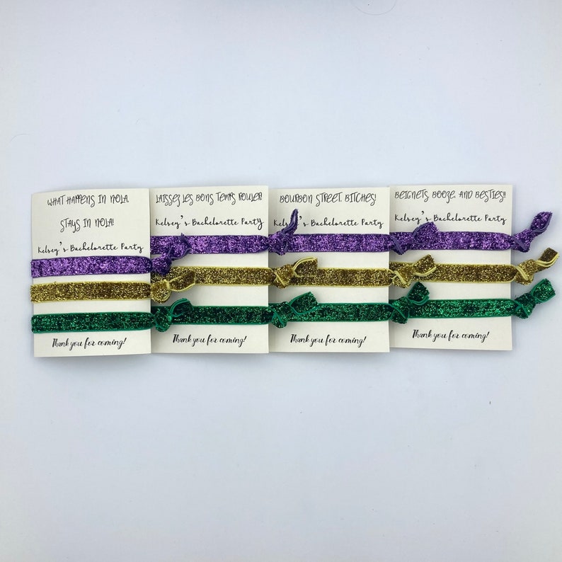 Peut inclure: Quatre cartes avec des dictons diff&eacute;rents li&eacute;s &agrave; la Nouvelle-Orl&eacute;ans, chacune avec trois &eacute;lastiques pour cheveux en violet, or et vert. Les dictons sont "What happens in NOLA stays in NOLA", "Laissez les bons temps rouler", "Bourbon Street Bitches", et "Beignets, booze, and besties". Chaque carte dit "Kelsey's Bachelorette Party" et "Thank you for coming!"