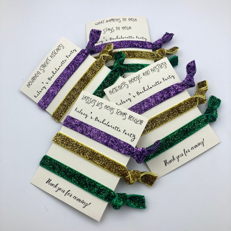 Peut inclure: Un ensemble de six &eacute;lastiques &agrave; cheveux en paillettes vertes, dor&eacute;es et violettes avec des cartes attach&eacute;es. Les cartes portent le texte suivant : "Bourbon Street Bitches ! Kelsey's Bachelorette Party", "What Happens in NOLA Stays in NOLA", "Beignets Booze, and Besties ! Kelsey's Bachelorette Party", "Laissez Les Bons Temps Rouler Kelsey's Bachelorette Party", et "Thank you for coming !"