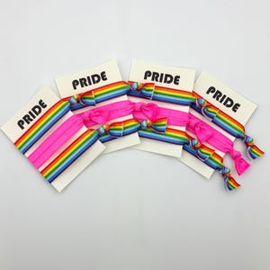 Könnte beinhalten: Eine Sammlung pinkfarbener Haargummis mit Regenbogenstreifen und Schleifen, jeweils befestigt an einer weißen Karte mit dem schwarzen Aufdruck "PRIDE". Die Haargummis sind fächerförmig vor weißem Hintergrund angeordnet.