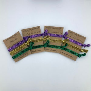 Peut inclure: Ensemble de six &eacute;lastiques &agrave; cheveux paillet&eacute;s violets, dor&eacute;s et verts avec des cartes de remerciement. Les cartes comportent diff&eacute;rentes phrases li&eacute;es &agrave; un enterrement de vie de jeune fille, notamment "What happens in NOLA stays in NOLA", "Bourbon Street Bitches" et "Betonets Booze and Besties".