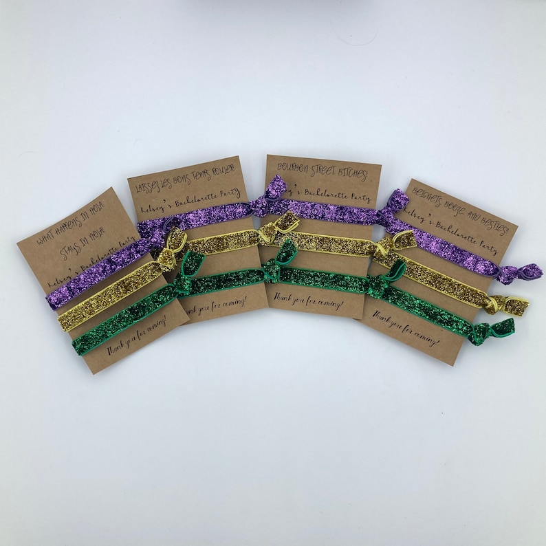 Peut inclure: Ensemble de six &eacute;lastiques &agrave; cheveux paillet&eacute;s violets, dor&eacute;s et verts avec des cartes de remerciement. Les cartes comportent diff&eacute;rentes phrases li&eacute;es &agrave; un enterrement de vie de jeune fille, notamment "What happens in NOLA stays in NOLA", "Bourbon Street Bitches" et "Betonets Booze and Besties".