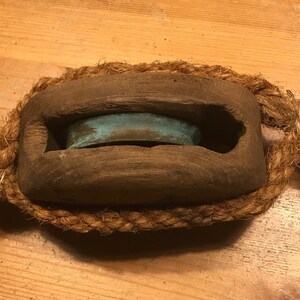 May include: A wooden pulley with a turquoise metal wheel, wrapped in natural rope with a black metal loop on each end.