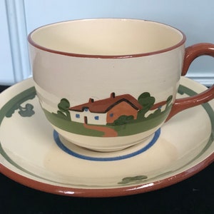May include: A cream-coloured teacup and saucer with a brown rim and a painted scene of a house with green trees and a path. The house has a brown roof, white walls, and blue windows.