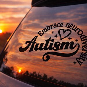 Könnte beinhalten: Schwarzer Aufkleber auf einer Autoscheibe mit dem Schriftzug "Embrace neurodiversity Autism" in einer stilisierten Schriftart, mit einem Herz und Sternen. Der Hintergrund ist ein Sonnenuntergang in Orange- und Gelbtönen.