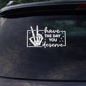 Könnte beinhalten: Weißer Aufkleber für die Heckscheibe eines Autos. Er zeigt eine Skeletthand, die ein Friedenszeichen macht, mit dem Text "have the day you deserve" in einer stilisierten Schriftart. Der Text ist in einem weißen rechteckigen Rahmen mit Sternen.