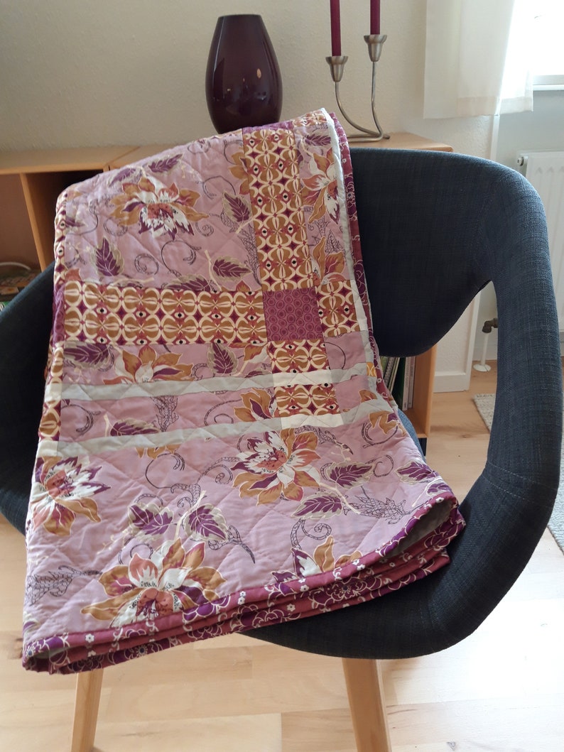 Puede incluir: Una colcha acolchada con un estampado floral en tonos rosa, marr&oacute;n y morado est&aacute; sobre una silla gris. La colcha presenta un dise&ntilde;o de patchwork con varios motivos geom&eacute;tricos y florales. Los bordes est&aacute;n acabados con un borde morado oscuro.