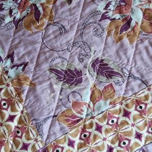 Puede incluir: Primer plano de una tela acolchada con un estampado floral. El dise&ntilde;o presenta grandes flores en tonos marr&oacute;n, rosa y blanco, sobre un fondo lila claro. El acolchado crea un patr&oacute;n diagonal en la tela.