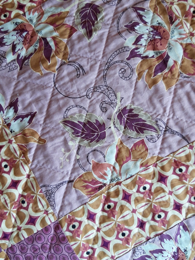 Puede incluir: Primer plano de una tela acolchada con un estampado floral. El dise&ntilde;o presenta grandes flores en tonos marr&oacute;n, rosa y blanco, sobre un fondo lila claro. El acolchado crea un patr&oacute;n diagonal en la tela.