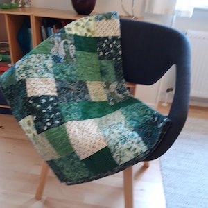 Może przedstawiać: Patchworkowa kołdra w odcieniach zieleni z kwiatowymi i paisley wzorami, narzucona na ciemnoszary fotel. Fotel ma drewniane nogi. W tle widoczny jest ciemnofioletowy wazon i świecznik.