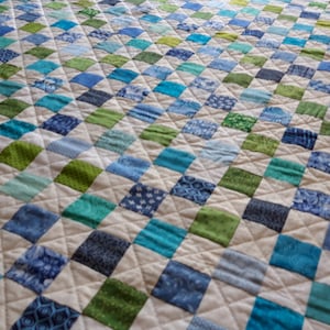 May include: A handmade quilt with a repeating pattern of blue and green squares on a cream background. The squares are various shades of blue and green, with some featuring patterned fabrics. The quilt is machine-stitched with a diamond pattern.