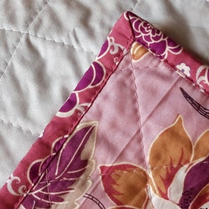 Puede incluir: Primer plano de una esquina de tela acolchada. La tela presenta un estampado floral en tonos rosa, morado y naranja. El borde est&aacute; rematado con un borde estampado en colores coordinados. El acolchado crea un patr&oacute;n de diamantes.