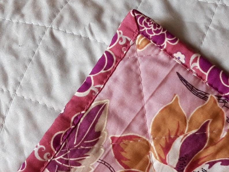 Puede incluir: Primer plano de una esquina de tela acolchada. La tela presenta un estampado floral en tonos rosa, morado y naranja. El borde est&aacute; rematado con un borde estampado en colores coordinados. El acolchado crea un patr&oacute;n de diamantes.