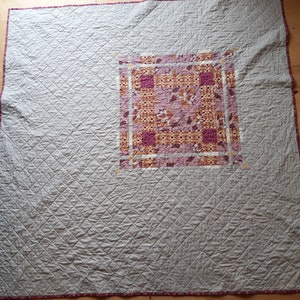 Puede incluir: Un edred&oacute;n hecho a mano con un cuadrado central de tela estampada en tonos rosa, naranja y blanco. El cuerpo principal del edred&oacute;n es de color gris claro con un patr&oacute;n de diamantes. Los bordes est&aacute;n ribeteados con un borde estampado.