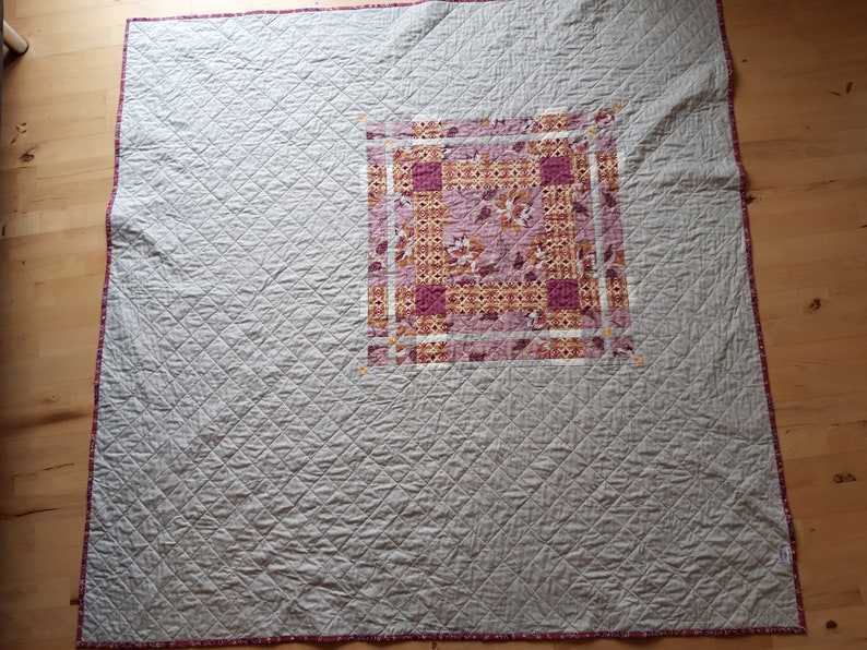 Puede incluir: Un edred&oacute;n hecho a mano con un cuadrado central de tela estampada en tonos rosa, naranja y blanco. El cuerpo principal del edred&oacute;n es de color gris claro con un patr&oacute;n de diamantes. Los bordes est&aacute;n ribeteados con un borde estampado.