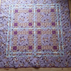 Puede incluir: Una colcha acolchada con un estampado floral en tonos morados, marrones y crema. La colcha presenta un dise&ntilde;o de cuadr&iacute;cula con bloques cuadrados y rectangulares. Los bordes est&aacute;n acabados con un borde azul claro.