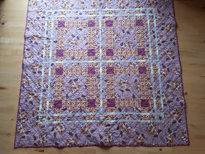 Puede incluir: Una colcha acolchada con un estampado floral en tonos morados, marrones y crema. La colcha presenta un dise&ntilde;o de cuadr&iacute;cula con bloques cuadrados y rectangulares. Los bordes est&aacute;n acabados con un borde azul claro.