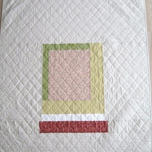 Może przedstawiać: Pikowany koc z kremowym tłem i centralnym wzorem. Wzór przedstawia kwadrat z kwiatowym wzorem, obramowany zielonymi i żółtymi obwódkami oraz czerwono-białym paskiem na dole. Koc ma wzór w romby.