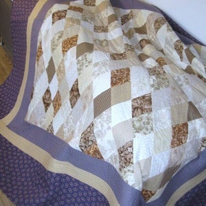 May include: A patchwork quilt in neutral tones. The central design is a mix of diamond and square patterns in white, beige, and brown. The border is a deep purple with a repeating pattern, and a tan accent. A cozy home decor item.