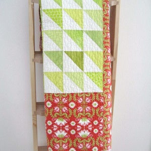 Può includere: Una trapunta fatta a mano con un design geometrico di triangoli verdi e bianchi nella metà superiore. La metà inferiore presenta un motivo floreale rosso con accenti bianchi e verdi. La trapunta è drappeggiata su una scala di legno.